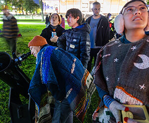 People gathered on the University Library Lawn at night look through a telescope; a person peers into it, while others look up at the sky.