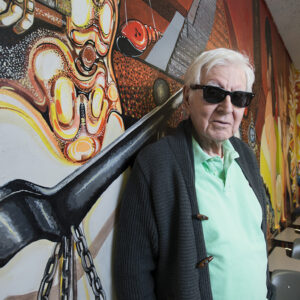 Rodolfo "Rudy" Acuña, wearing sugnlasses, stands in front of a brightly colored mural.