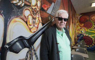 Rodolfo "Rudy" Acuña, wearing sugnlasses, stands in front of a brightly colored mural.