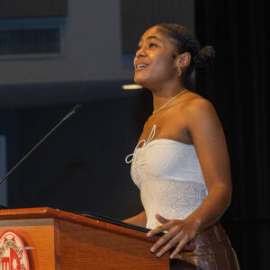 A CSUN student in a white halter top stands, singing at a wooden podium. She is looking up into the distance.