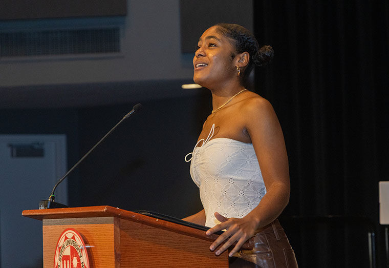 A CSUN student in a white halter top stands, singing at a wooden podium. She is looking up into the distance.