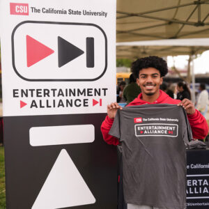 Student holding a CSU Entertainment Alliance t-shirt next to a CSU Entertainment Alliance sign.