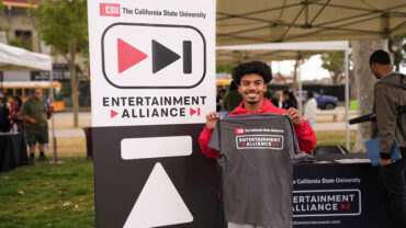 Student holding a CSU Entertainment Alliance t-shirt next to a CSU Entertainment Alliance sign.