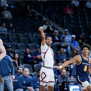 A male basketball player and two defenders watch after he releases a 3-point shot.
