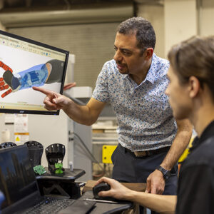 Peter Bishay gestures toward a computer screen displaying a robotic arm, while a student, seated, looks on.