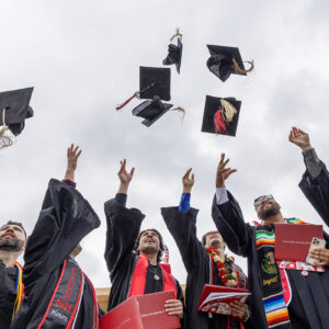 Four graduates in black robes and colorful sashes toss their graduation caps in the air.