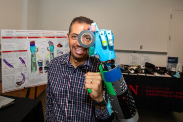 Peter Bishay uses a brightly colored prosthetic arm and hand to hold a roll of tape up to his eye.