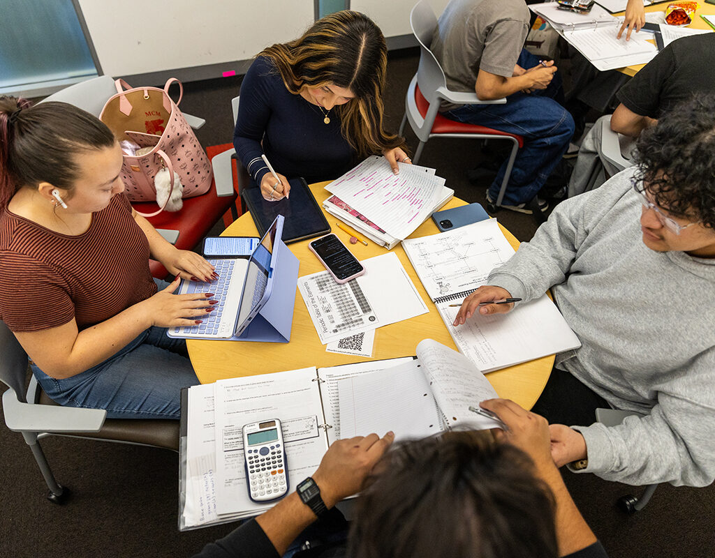 A group of students sit around a table with notebooks and papers spread in front of them.