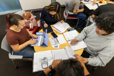 A group of students sit around a table with notebooks and papers spread in front of them.