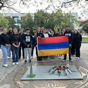 A group of Armenian students hold an Armenian flag in front of a wooden replica of an Armenian Genocide memorial eternal flame. They are standing in Matador Square.