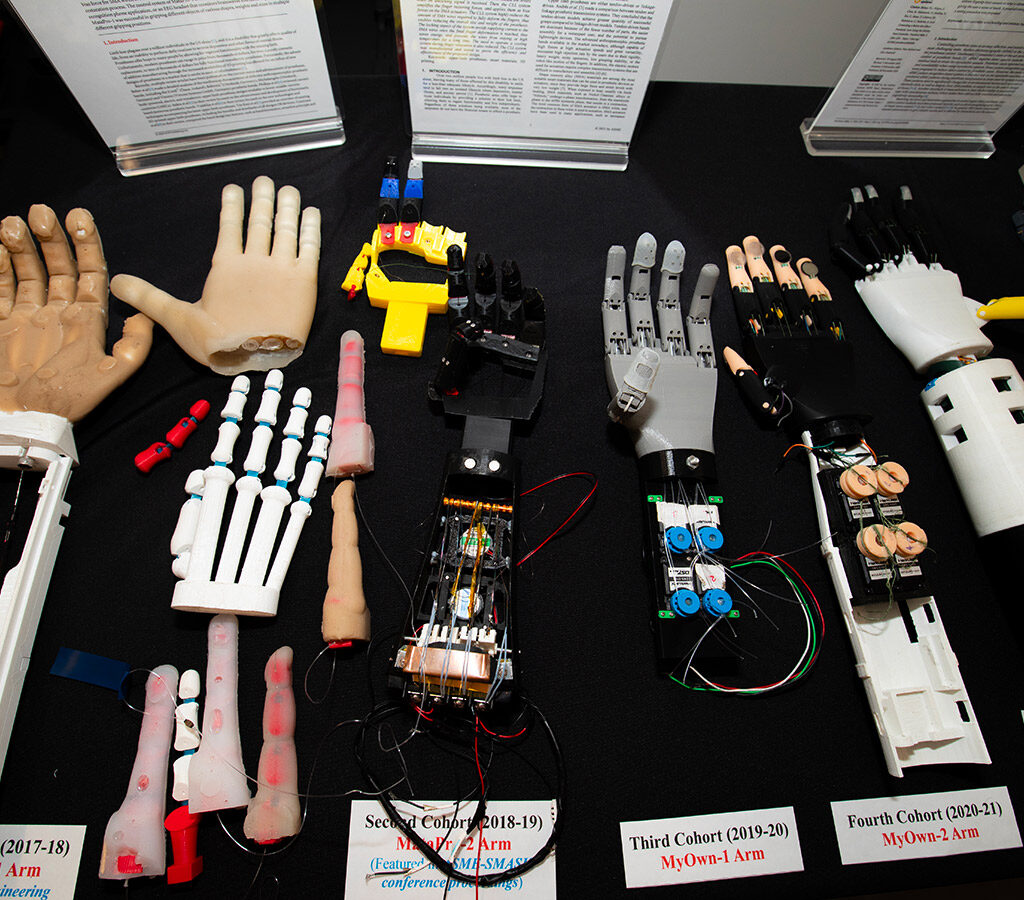 Five prosthetic arms with their wiring exposed, a prosthetic hand and prosthetic fingers are lined up on a table, with labels of each student cohort's work.