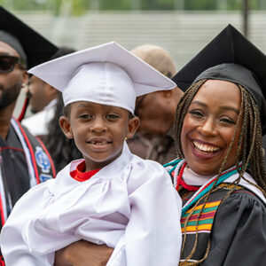 A smiling graduate in a cap and gown holds a child wearing a white graduation cap and gown at Black Graduation.