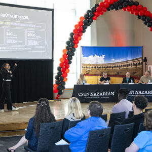Person stands on stage, gesturing to a large video screen, while a panel of four judges, seated, looks on.