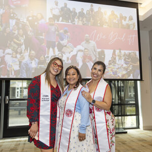 Three women wearing white sashes pose for a photo in front of a large video screen.