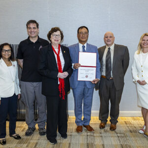 Six people stand together for a photo. The man in the center holds a certificate.