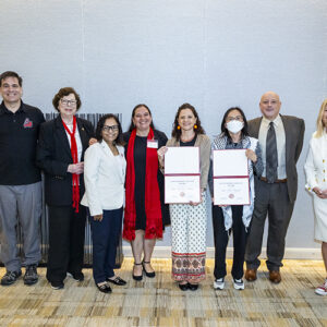 Eight people stand together, with the two people in the center holding up certificates