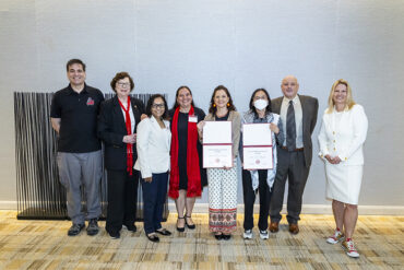 Eight people stand together, with the two people in the center holding up certificates