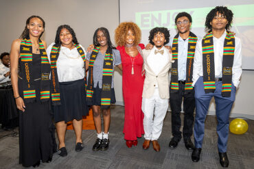 Students wearing colorful stoles and Theresa White stand together with their arms around each other.