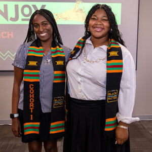Two students wearing colorful graduation stoles stand together, smiling.