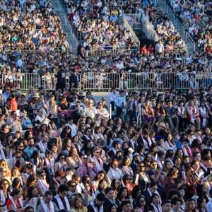 A large crowd of students seated in chairs on the grass in the foreground, friends and family members in the stands behind them.
