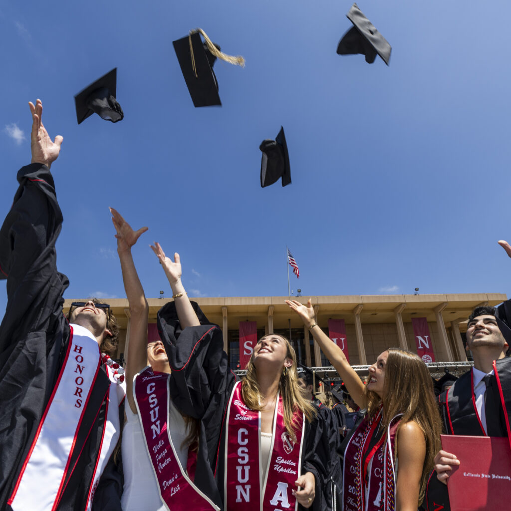 Five graduates toss their caps high in the air against a blue sky in front of the CSUN library.