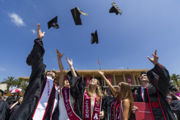 Five graduates toss their caps high in the air against a blue sky in front of the CSUN library.