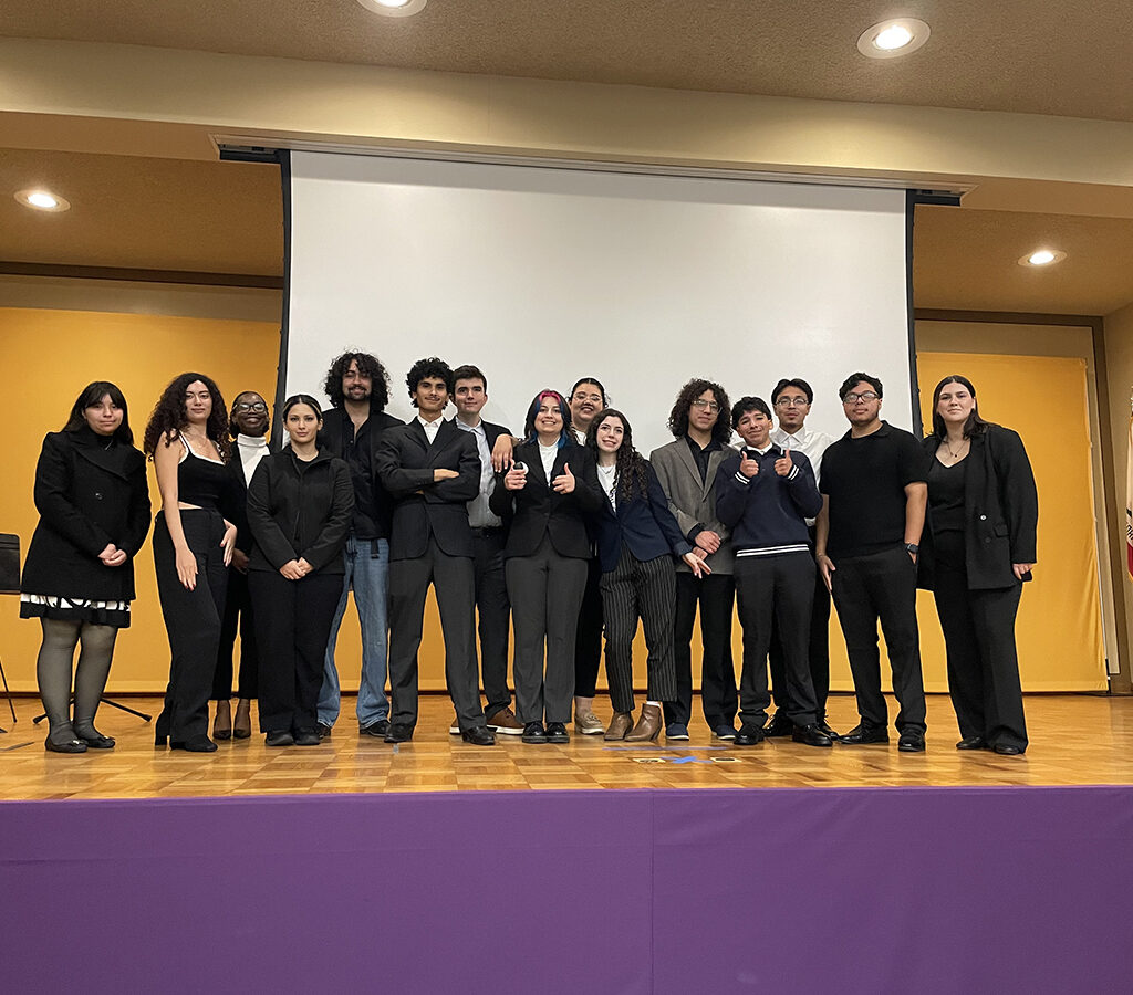 A group of 15 students stands on a stage witha video screen behind them.