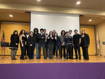 A group of 15 students stands on a stage witha video screen behind them.
