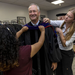 Two people assist Doug Emhoff with his academic regalia before heading to the commencement stage.