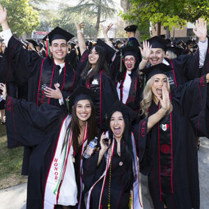 A group of graduates in caps and gowns stand with their hands in the air, cheering. Three are kneeling down in front. A crowd of other graduates line up behind them on a grassy lawn.