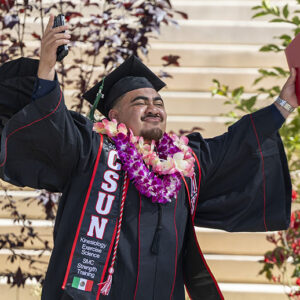 A graduate in cap and gown holds a red diploma cover in the air.