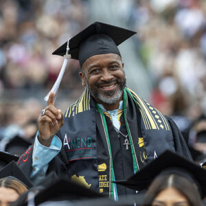 A CSUN graduate in cap and gown stands up in the crowd of graduates, pointing his index finger in the air and smiling.
