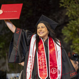 A CSUN graduate in cap and gown walks across the Commencement stage, holding a red diploma cover in the air.