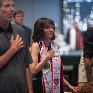 A woman in a white and red graduation sash stands with her right hand over her heart.