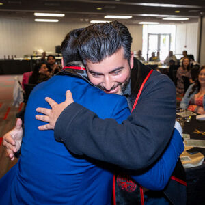 Two men hug, with the man facing the camera smiling. Both are wearing black and red graduate sashes.
