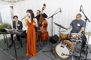 A woman in a long gown stands at a microphone with musicians in the background.