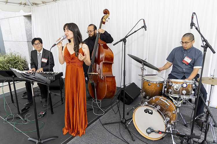 A woman in a long gown stands at a microphone with musicians in the background.
