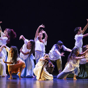 Students wearing white shirts dance on stage.