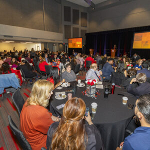 People seated at tables in a large room, with video screens in the background.