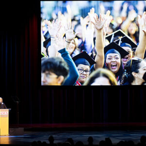 CSUN President Erika D. Beck stands at a podium on stage next to a large video screen displaying a photo of graduates.