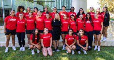 Student athletes dressed in red shirts pose for a group photo on the grass with CSUN President Erika D. Beck.