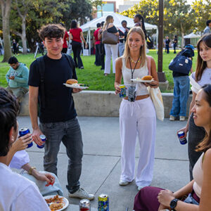 Students holding plates of food stand together on the sidewalk.