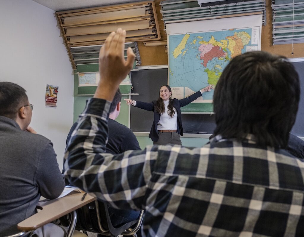 A female history teacher points to a map while a student raises his hand.