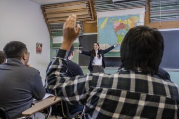 A female history teacher points to a map while a student raises his hand.
