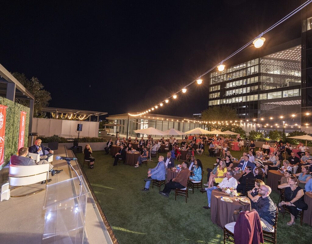 A packed CSUN alumni event with Doug Emhoff on stage on a brightly lit LA rooftop at night. 